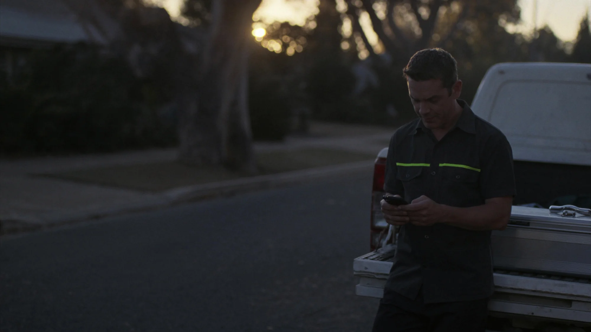 Sample photo: a tradesman in a work shirt with a hi-vis stripe checks his phone beside a ute at dawn